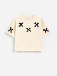 T-shirt z kokardkami - kremowy. Białe koszulki dziewczęce Reserved, bez wzorów, z bawełny, bez ramiączek. Za 39.99 zł.